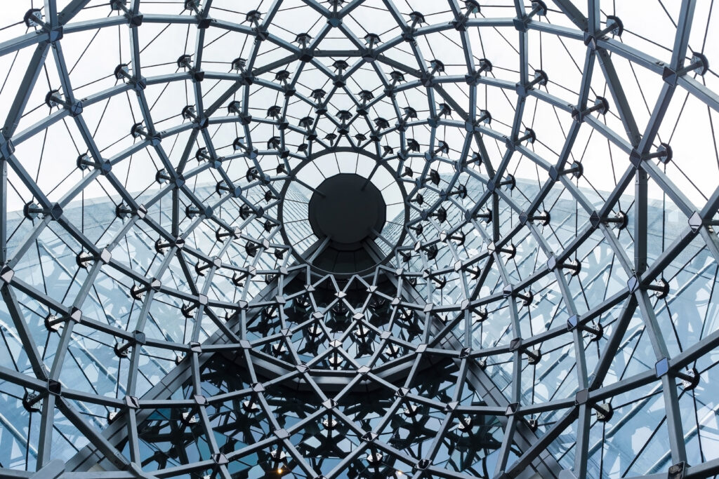 Zicht van onderaf op een grote glazen koepel met een symmetrisch stalen raamwerk en driehoekige panelen rond een centrale ronde opening in grijsblauwe tinten.