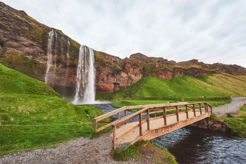 Een waterval valt langs een rotswand met een houten brug over de rivier in een groen IJslands landschap.