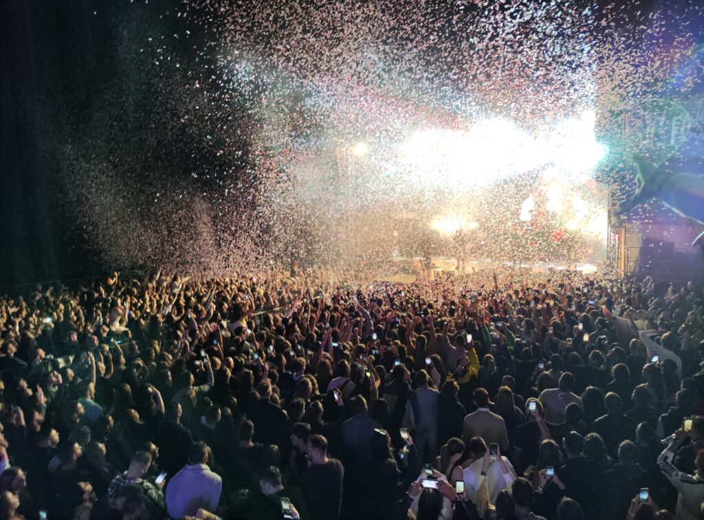 Confettiregen boven een grote concertmenigte voor een fel verlicht podium.